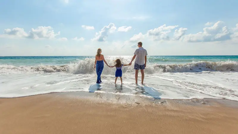 Family holding hands facing the sea on a Bali beach, waves crashing at their feet.