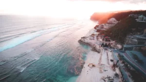 Aerial view of Melasti Beach Ungasan, Bali, showcasing turquoise waters, white sand, and dramatic limestone cliffs at sunset.