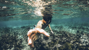 Snorkeller exploring clear waters of Bali, reaching out towards the camera with coral seabed in the background.