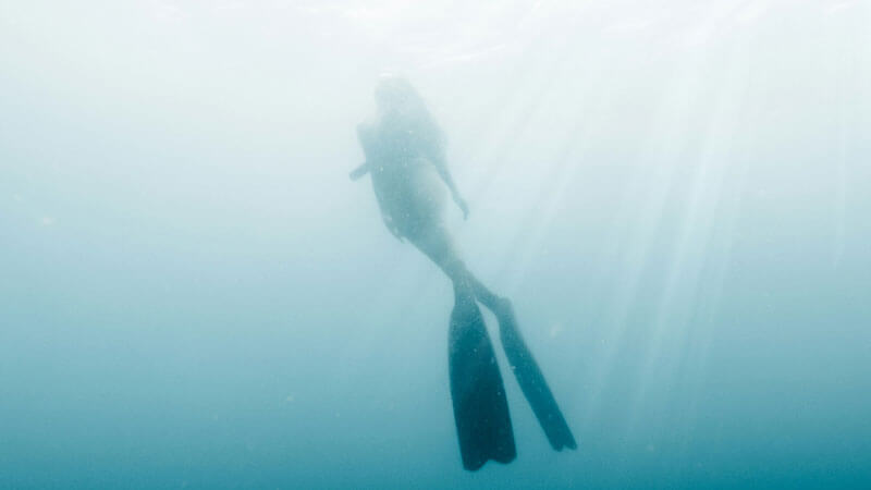 Silhouette of a freediver exploring the tranquil underwater world off Amed Beach in East Bali, with sunbeams piercing through the ocean surface.