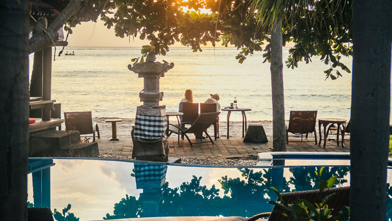 Couple enjoying a serene sunset from a beachfront cafe near Amed, Bali, with traditional Balinese décor and a tranquil sea view.