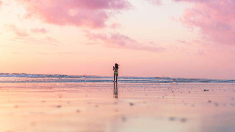 Silhouette of a woman walking on Bali beach at sunset with a pink sky.