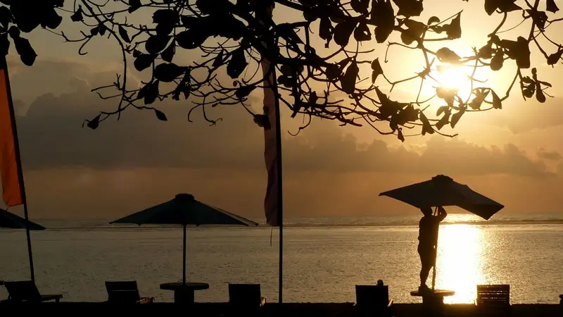 Beachfront dining setup in Sanur with chairs and umbrellas silhouetted.