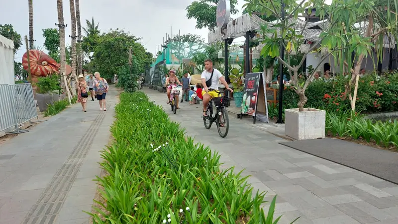 Cyclists and pedestrians enjoying the scenic Sanur Promenade with lush greenery and beachfront cafés.