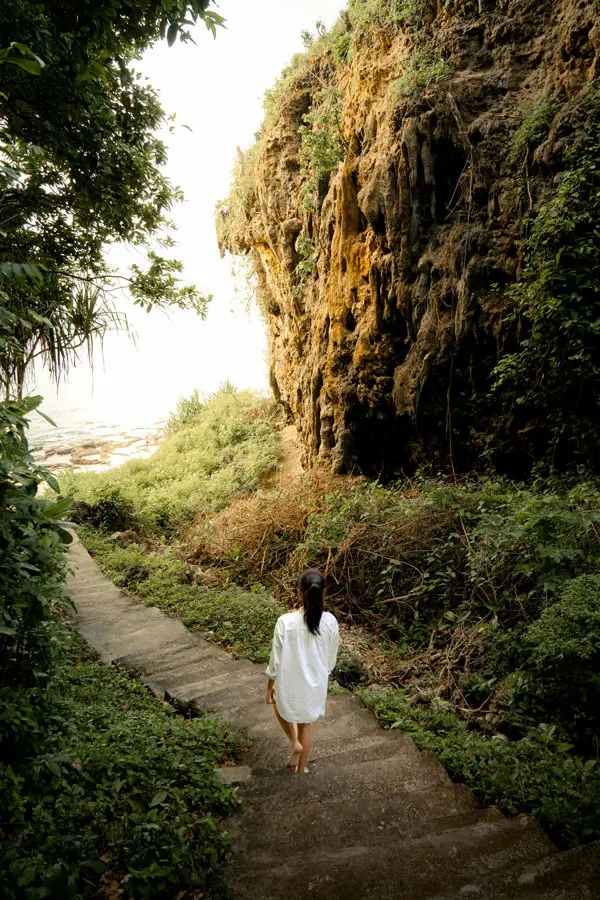 Solo traveler walking down a natural cliffside trail surrounded by tropical plants and limestone rock.