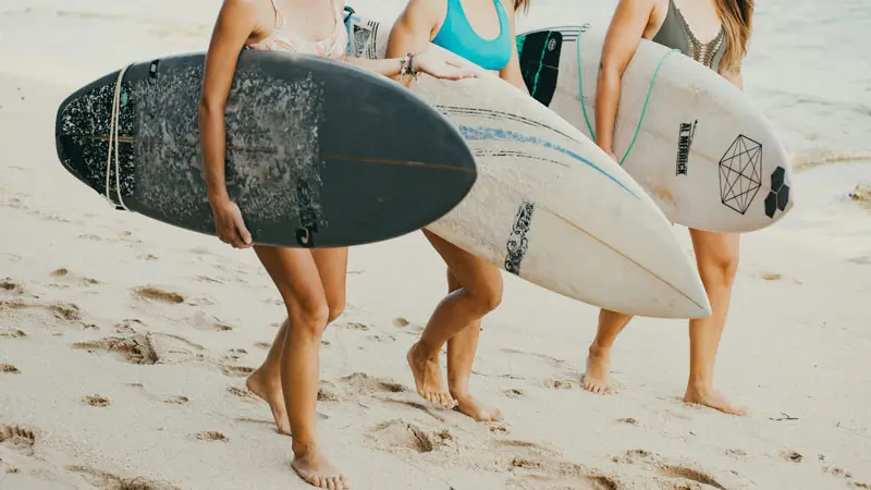 Women surfers walking on sandy beach carrying shortboards together.