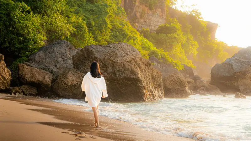 Solo female traveler walking barefoot at Balangan Beach Bali during golden hour with cliffs in the background.