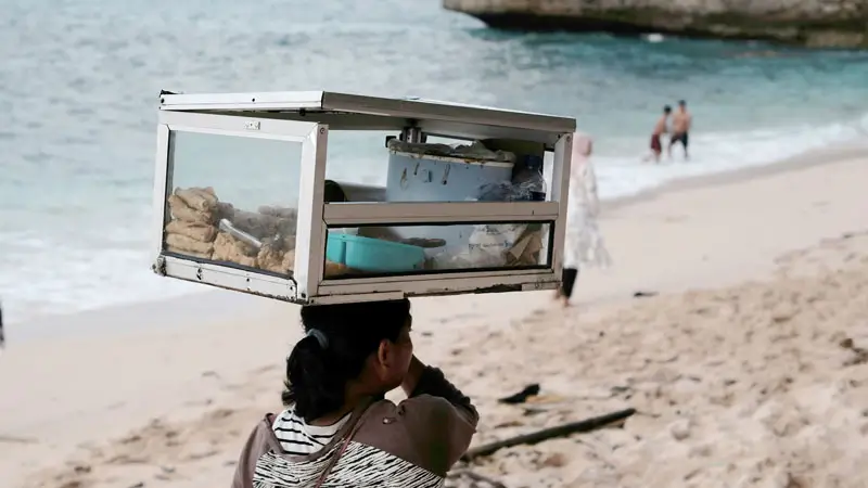 Local snack vendor walking along a tropical beach with food case.