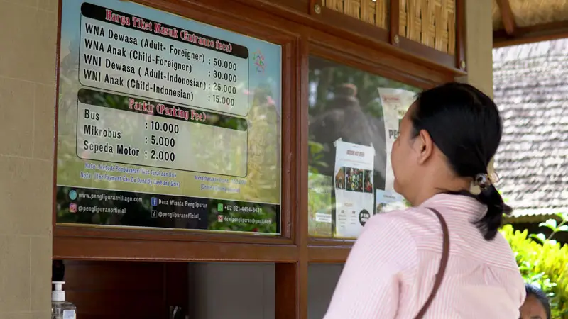 Visitor buying ticket at Penglipuran village counter; board shows WNI 25,000 and WNA 50,000