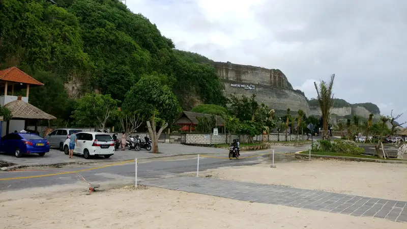 Beach-level parking area at Melasti Beach with cars and scooters below cliffs.