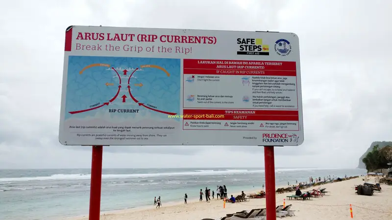 Rip current warning sign at Melasti Beach with safety instructions on the sand.