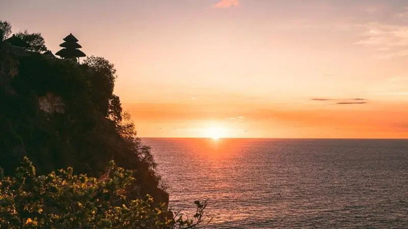 Uluwatu cliff at sunset with temple silhouette above the ocean.