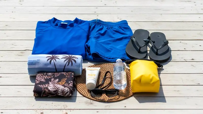 Flat lay of swimwear, towel, sunscreen, water, flip-flops, and a small dry bag.